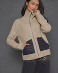 A woman with long dark hair is posing against a gray background, wearing the Deep Pockets Zip by Lisa Todd. This textured stitch cotton blend zip-up sweater features a high collar and two black patch pockets. She pairs it with dark pants and rests one hand on her chin.