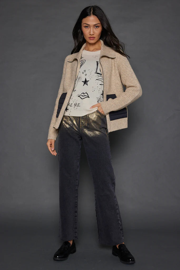 A woman with long dark hair is positioned against a gray backdrop, showcasing the Lisa Todd Deep Pockets Zip jacket in beige knit over a graphic tee that displays abstract face designs. She complements her outfit with dark metallic pants and black shoes, placing her hands in her pockets as she looks directly into the camera.