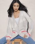 A person with long dark hair is sitting on a wooden stool, wearing the Lisa Todd "Camo Cloud" cardigan with contrast striping detail over a white top and blue jeans. The background is plain and neutral.