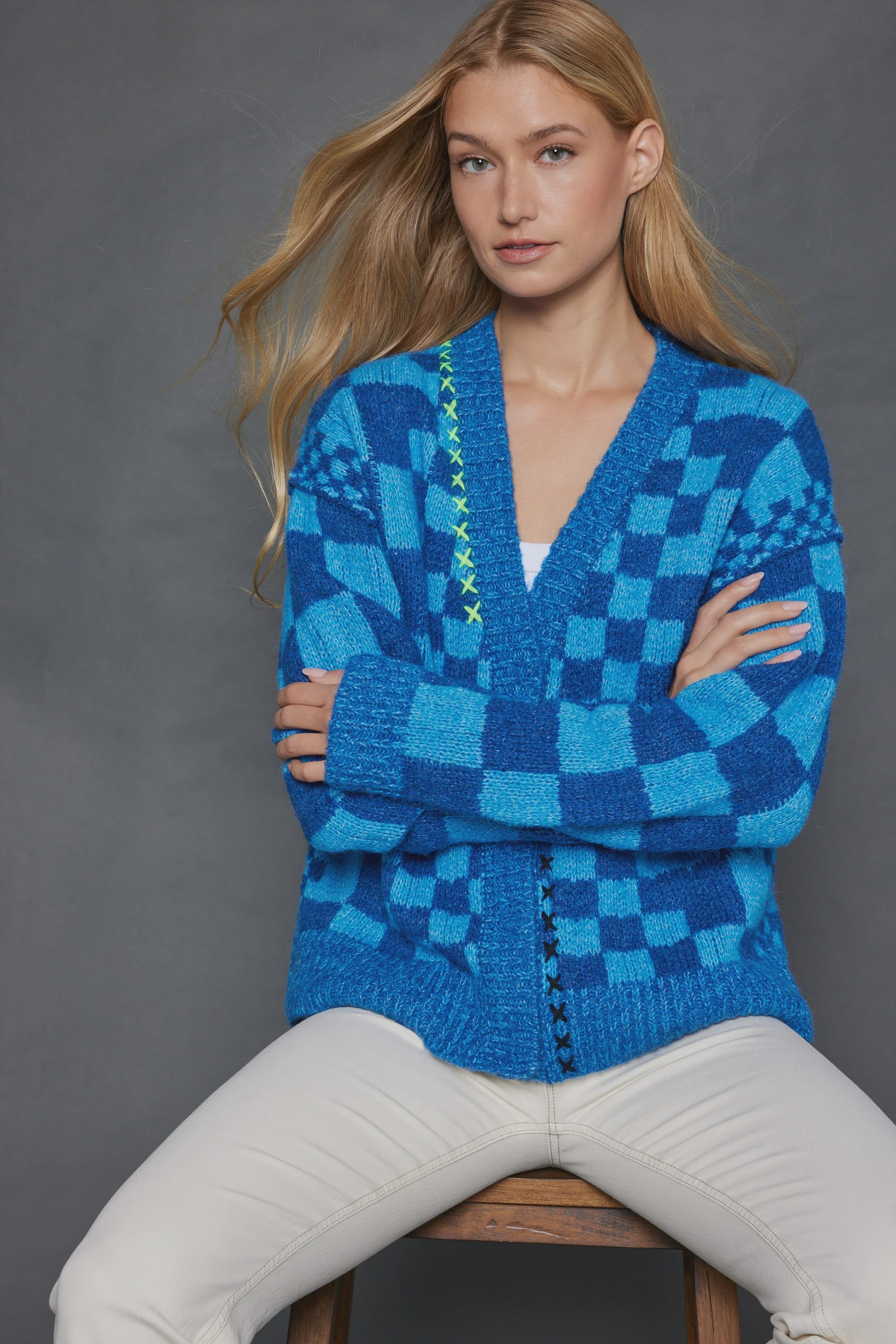 A person with long blond hair sits on a wooden stool, arms crossed. They wear Lisa Todd's "Check Me Out Card," a blue snap-front cardigan featuring light blue and white embroidery on one side, paired with white pants. The plain dark gray background highlights the relaxed fit of their checkered pattern ensemble.