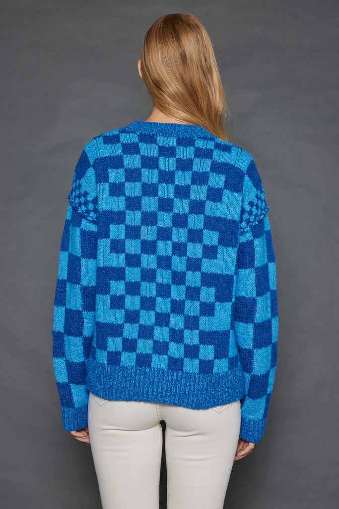 A person with long hair is seen from behind, dressed in the "Check Me Out Card" by Lisa Todd, a relaxed fit sweater featuring blue and turquoise checkered patterns with textured designs on the shoulders. Paired with light-colored pants, they stand against a plain gray background.