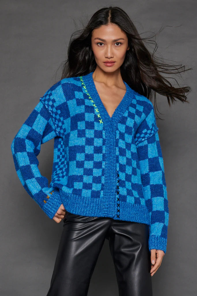 A person wearing the Check Me Out Card by Lisa Todd, featuring colorful details, poses in black leather pants against a gray background. The relaxed fit of the knit complements their long, dark hair flowing freely.