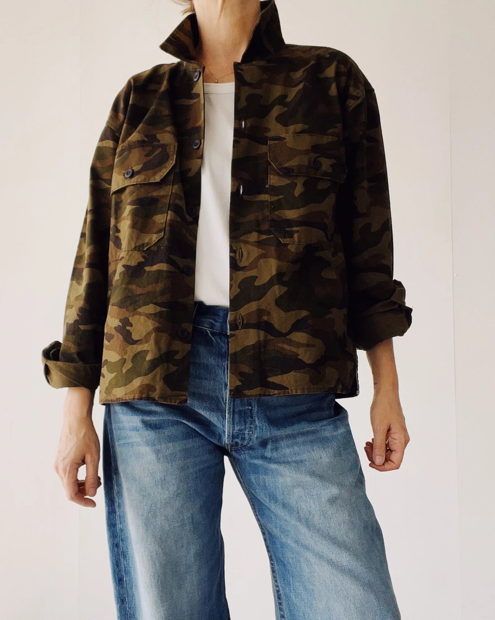 A person models The Work Shirt by Hey Gang—a boxy fit camouflage jacket styled over a white shirt and blue jeans—standing against a plain light background with their face not shown.