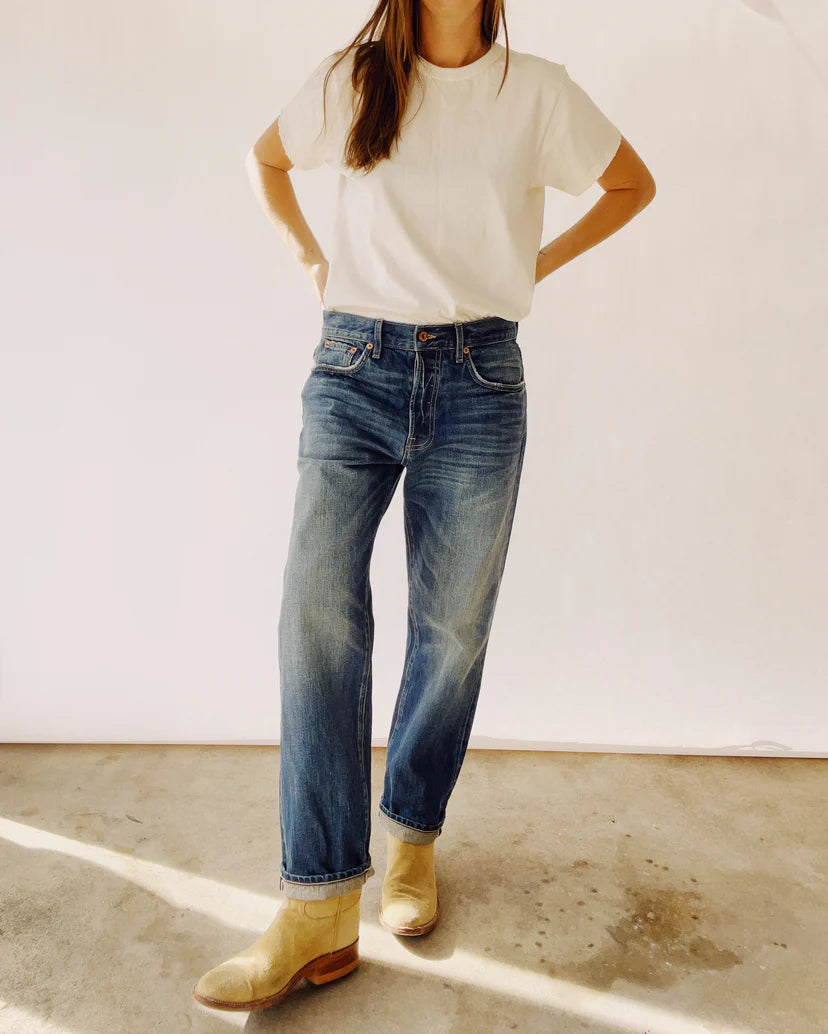 The individual stands casually on a concrete floor wearing "The Loose Crop Selvedge" high-rise jeans by Hey Gang, paired with a white t-shirt and light-colored boots. Their hands rest on their hips, casting a shadow on the ground, while the background remains simple and unobtrusive.