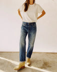 The individual stands casually on a concrete floor wearing "The Loose Crop Selvedge" high-rise jeans by Hey Gang, paired with a white t-shirt and light-colored boots. Their hands rest on their hips, casting a shadow on the ground, while the background remains simple and unobtrusive.