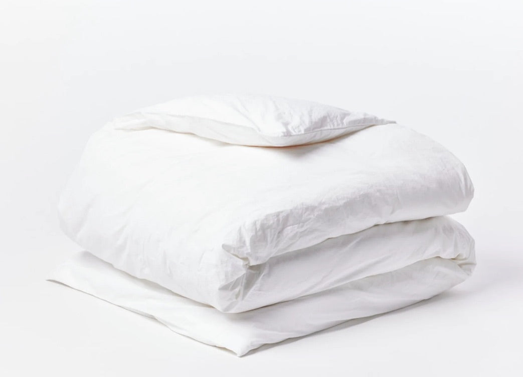 A neatly folded white duvet and pillow, crafted from GOTS certified organic cotton bedding, are placed against a plain white background, highlighting the crisp, clean fabric and soft texture. The simplicity and cleanliness of the Organic Crinkled Percale Duvet Alpine White Queen by Coyuchi Inc are emphasized in this minimalist arrangement.