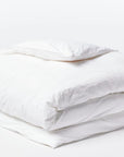 A neatly folded white duvet and pillow, crafted from GOTS certified organic cotton bedding, are placed against a plain white background, highlighting the crisp, clean fabric and soft texture. The simplicity and cleanliness of the Organic Crinkled Percale Duvet Alpine White Queen by Coyuchi Inc are emphasized in this minimalist arrangement.