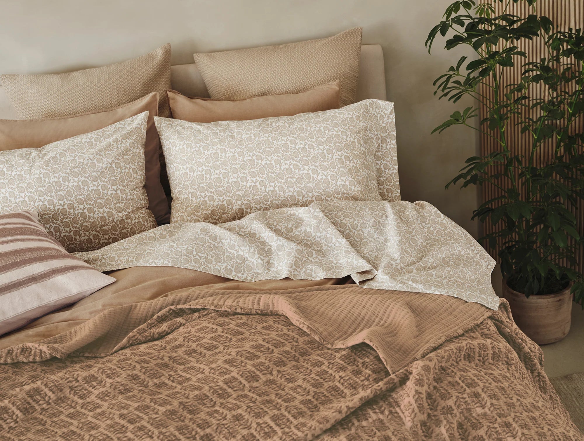 A neatly made bed features patterned beige and white pillows, a striped pillow, and beige and pink bedding. The Coyuchi Inc Mirada Matelasse Blanket in organic cotton adds comfort. A potted green plant sits by the bed against a light wall.