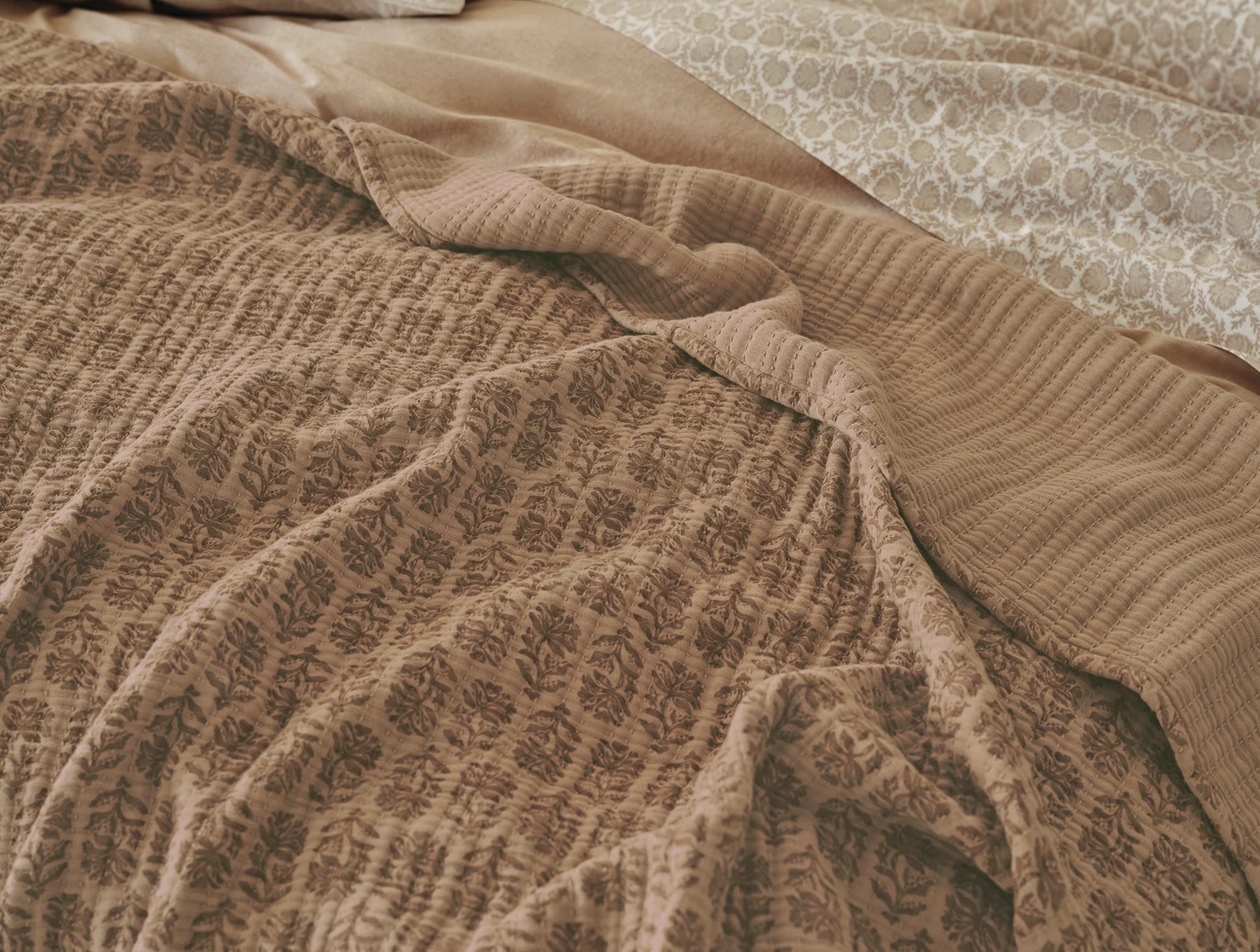 A close-up of Coyuchi Inc’s Mirada Matelasse Blanket shows beige and light brown jacquard fabric in subtle floral and geometric patterns, evoking a vintage Indian woodblock look with softly wrinkled, layered texture.