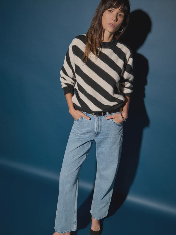 A woman wearing the Fabienne Stripe Sweater by Nation LTD and light blue wide-leg jeans stands against a dark blue backdrop, casting a shadow. With her long hair styled with bangs and hands casually tucked in her pockets, she exudes a stylish yet relaxed vibe.