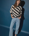 A woman wearing the Fabienne Stripe Sweater by Nation LTD and light blue wide-leg jeans stands against a dark blue backdrop, casting a shadow. With her long hair styled with bangs and hands casually tucked in her pockets, she exudes a stylish yet relaxed vibe.