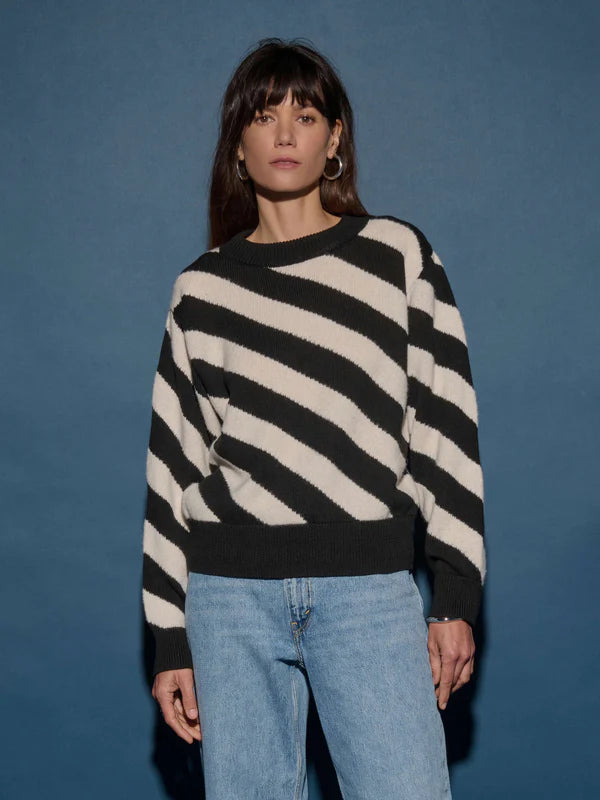 A person with long dark hair stands against a blue background, wearing the Fabienne Stripe Sweater by Nation LTD, featuring bold black and white diagonal stripes, paired with light blue jeans. They have a neutral expression and are looking at the camera.