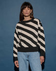 A person with long dark hair stands against a blue background, wearing the Fabienne Stripe Sweater by Nation LTD, featuring bold black and white diagonal stripes, paired with light blue jeans. They have a neutral expression and are looking at the camera.