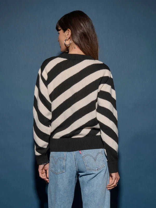 A person with long brown hair stands with their back to the camera, wearing the easy-fit Fabienne Stripe Sweater by Nation LTD, which features bold black and white diagonal stripes, paired with light blue jeans against a dark blue backdrop.