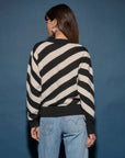 A person with long brown hair stands with their back to the camera, wearing the easy-fit Fabienne Stripe Sweater by Nation LTD, which features bold black and white diagonal stripes, paired with light blue jeans against a dark blue backdrop.