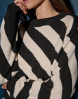 A person wearing the Fabienne Stripe Sweater by Nation LTD, featuring bold black and white stripes, gently rests their hand on their chin. The easy fit of the wide horizontal stripes, paired with the soft shadow effect from the lighting, perfectly complements the muted dark blue background.