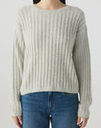 A woman wears the AMO Denim Hallie Sweater, a relaxed boxy knit in light gray alpaca wool with a ribbed crew neck, paired with blue jeans. She stands against a plain light background; the image is cropped above her head and at her thighs.