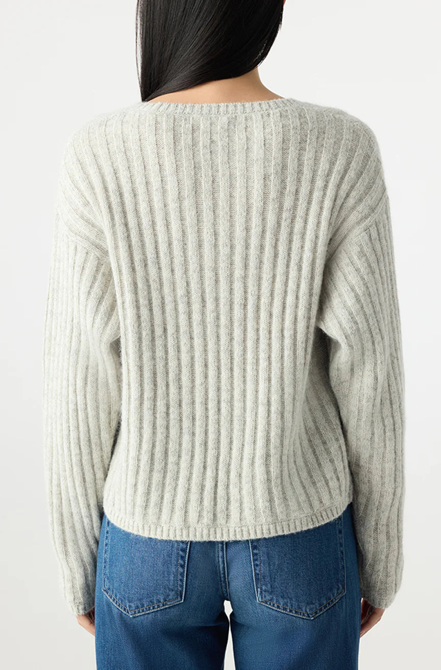 A person with long dark hair, seen from behind, wears the AMO Denim Hallie Sweater in light gray—a relaxed, boxy alpaca knit—paired with blue jeans against a plain light background.