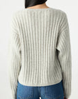 A person with long dark hair, seen from behind, wears the AMO Denim Hallie Sweater in light gray—a relaxed, boxy alpaca knit—paired with blue jeans against a plain light background.