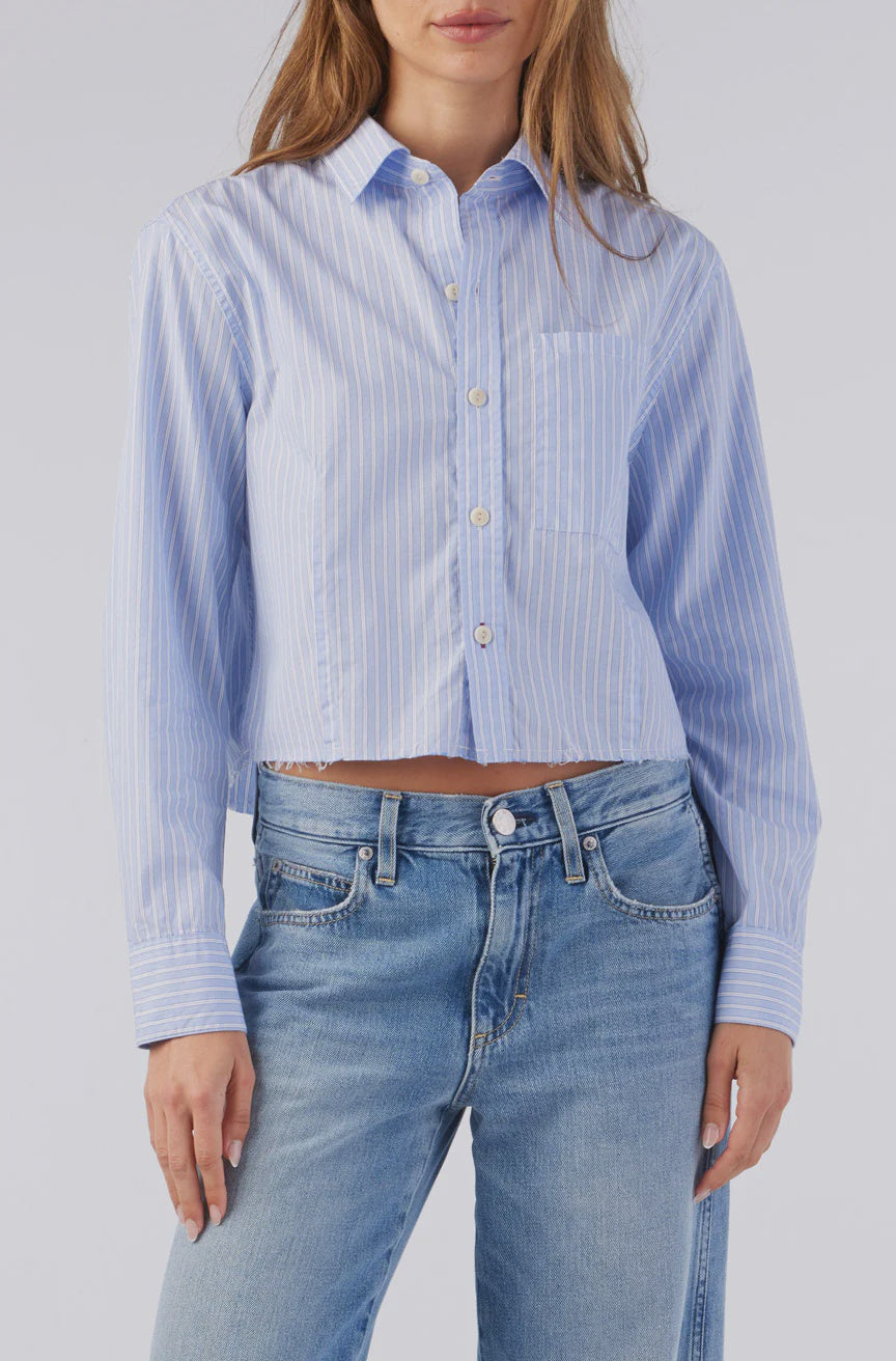 A person wearing AMO Denim's Selma Shirt, featuring a blue multi-stripe design with long sleeves and 100% cotton for a relaxed fit, tucked into light blue jeans. The shirt has a front pocket, and their loose hair partially covers their shoulders.