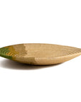 A Restorative Woven Bowl 21" Moss by Kazi Goods, a shallow, oval-shaped woven basket in natural tan color with subtle green accents radiating outward from the center is showcased against a plain white background. This piece reflects the tranquil aesthetic of a Scottsdale Arizona bungalow.