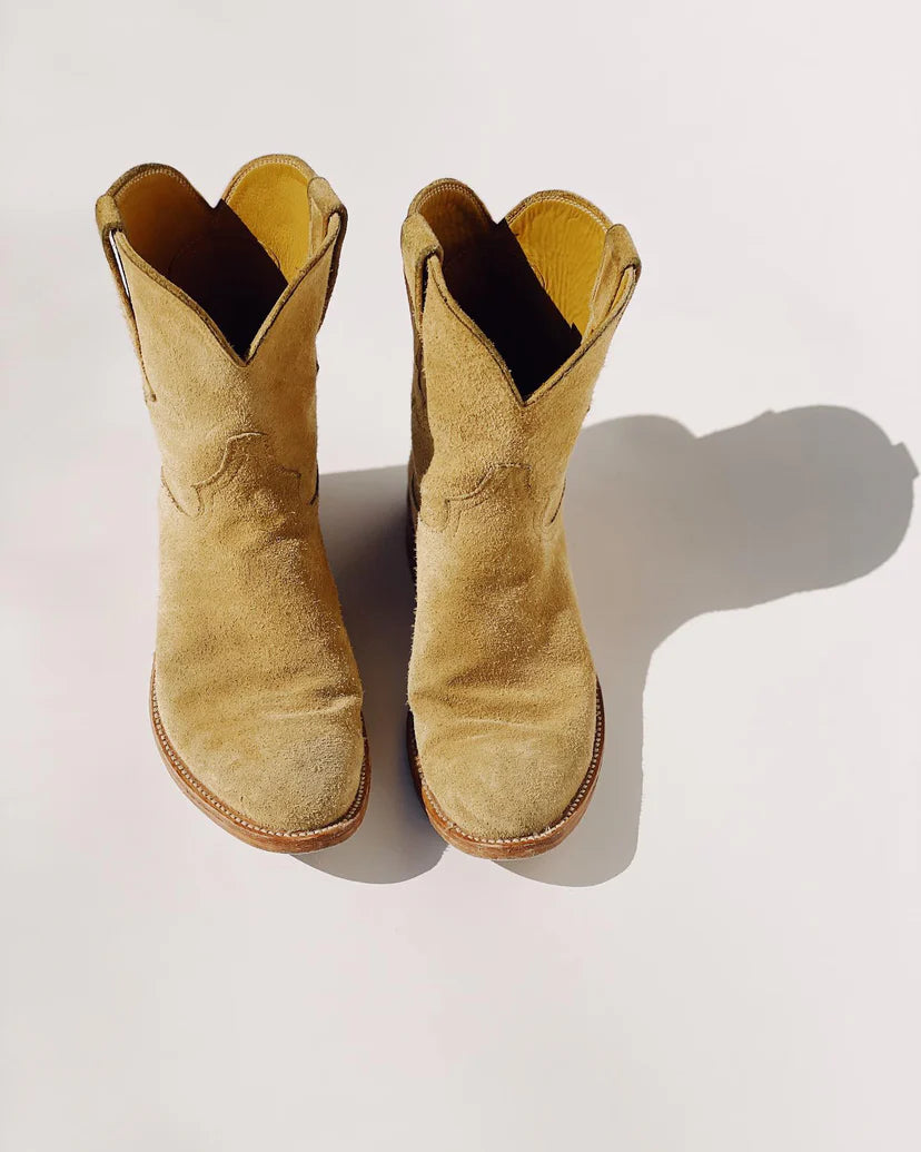 A pair of The Ranch Boot by Hey Gang, characterized by their roper style and crafted from tan calf skin rough out suede, showcase pointed toes and slightly curved tops. They are elegantly placed on a smooth white surface with sunlight casting a distinct shadow to the right.