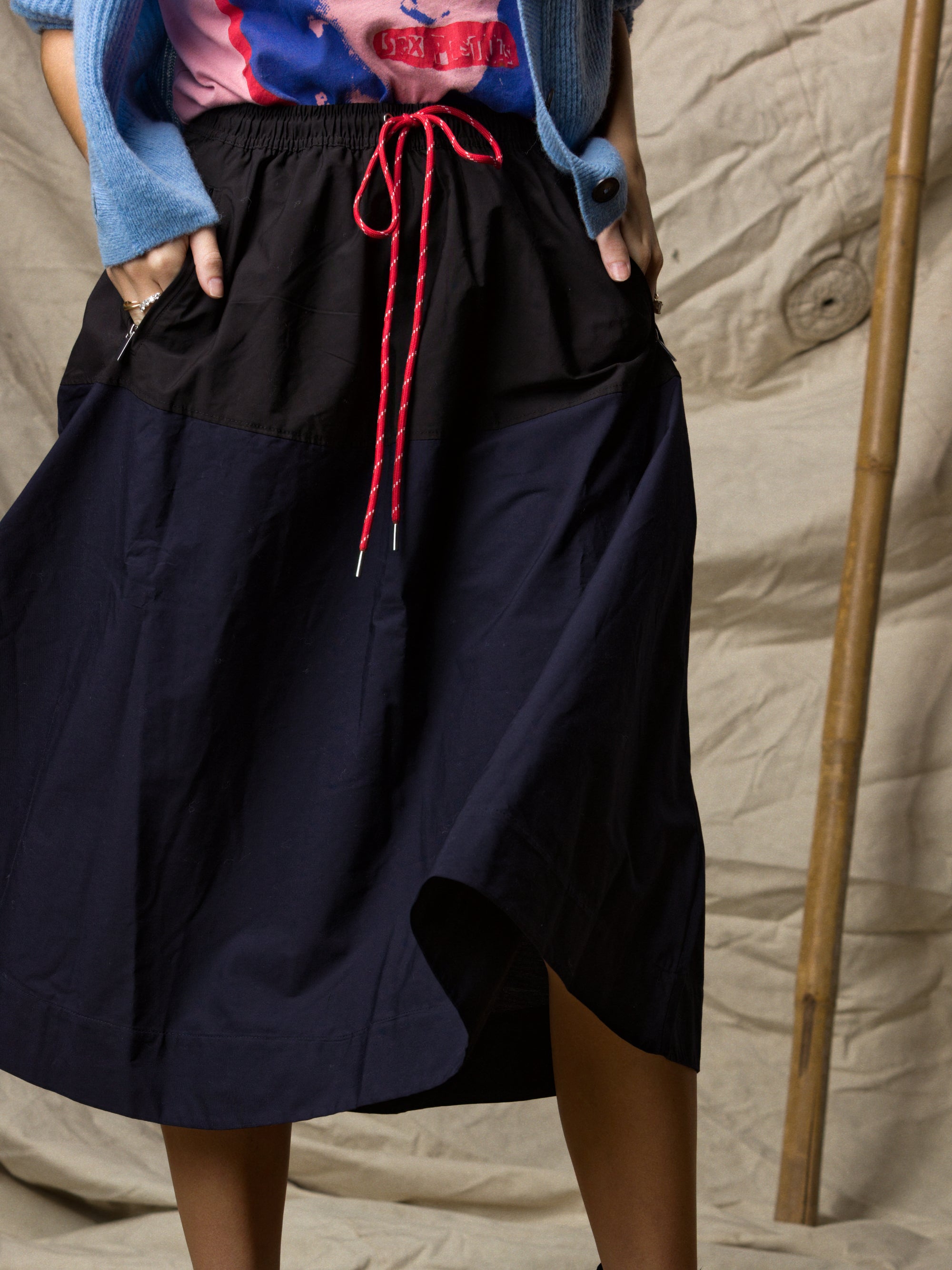 Someone wearing the Ping Pong Surf Club Le Vintage Sport Skirt in black with red drawstrings, paired with a blue cardigan and colorful top, stands before a beige textured background and bamboo pole. Face not shown.