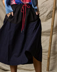 Someone wearing the Ping Pong Surf Club Le Vintage Sport Skirt in black with red drawstrings, paired with a blue cardigan and colorful top, stands before a beige textured background and bamboo pole. Face not shown.