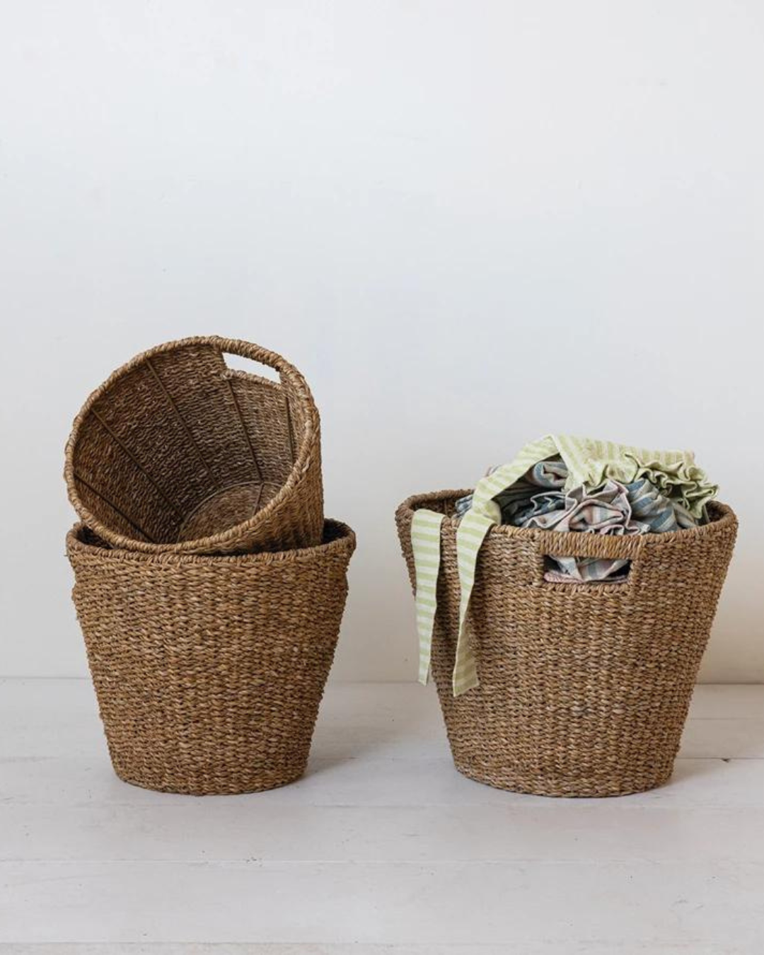 A still life image features two Round Woven Baskets by Creative Co-op placed against a plain white background. The basket on the left is empty with another basket nested inside it, showcasing their natural charm. The basket on the right contains rumpled, colorful fabric with green and white striped material on top, emphasizing an eco-friendly aesthetic.