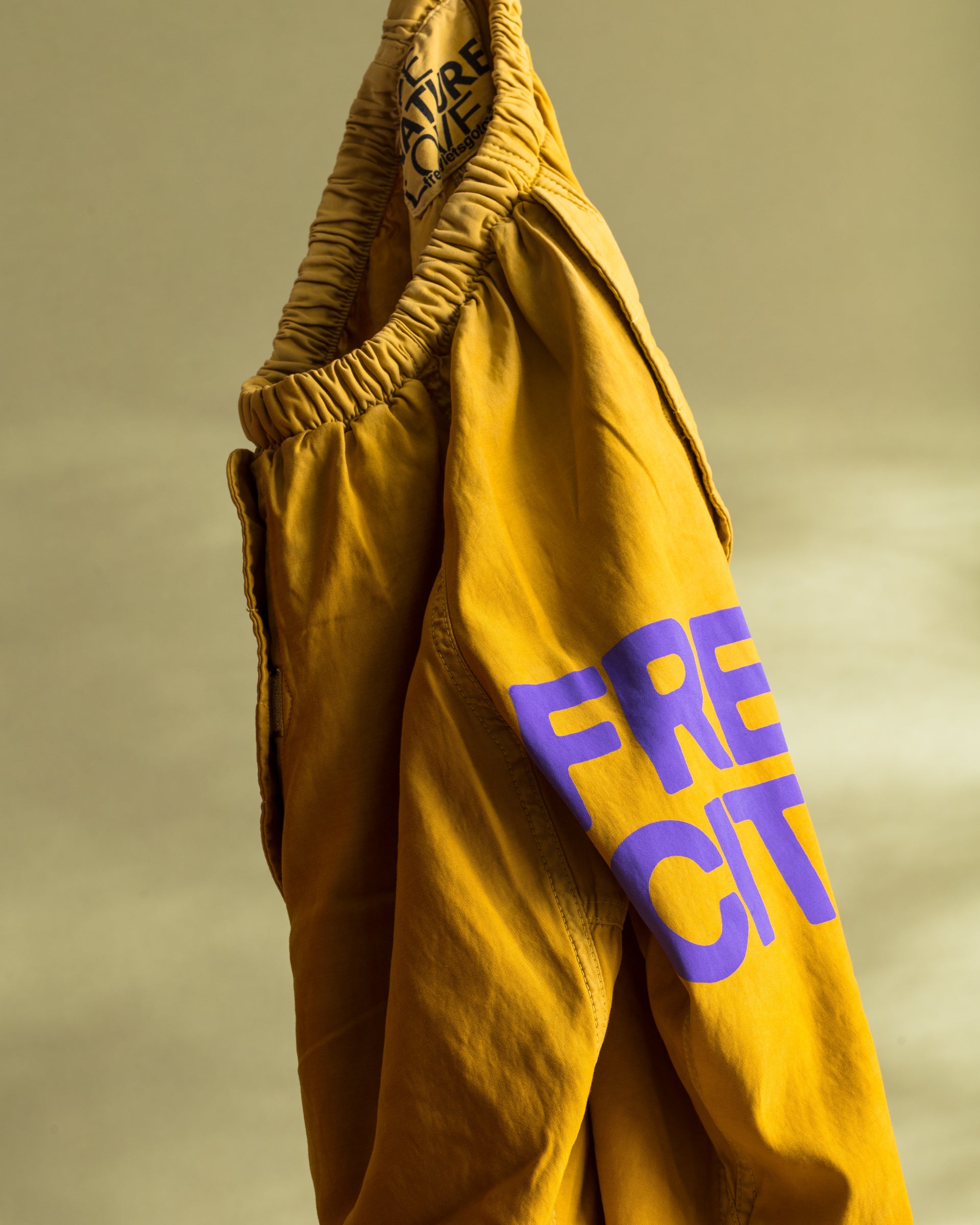 A pair of Flap/Snap LG MATTE/SATIN AirJump Pants in mustard-yellow, adorned with a purple &quot;FREE CITY&quot; logo on an elastic waistband, featuring a custom sunfade wash, set against a yellow-green background. 

Brand: Free City (sparrow, LLC)