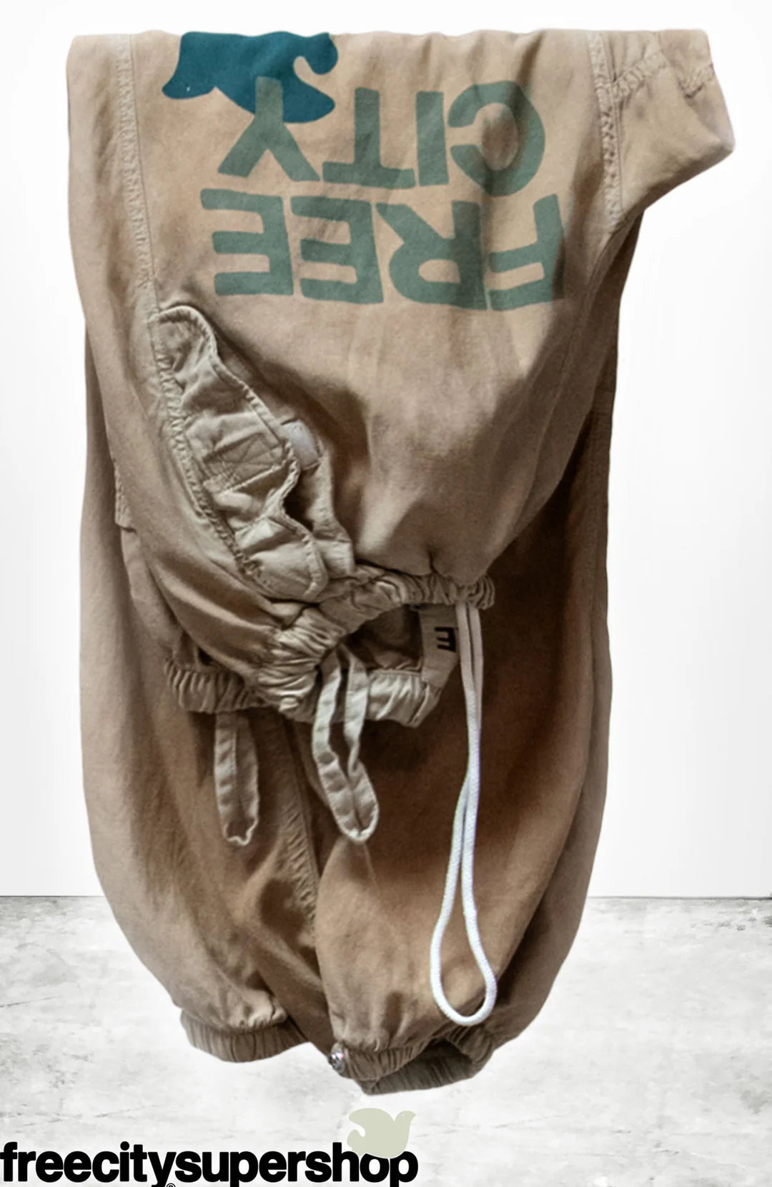 A pair of Flap/Snap LG MATTE/SATIN AirJump Pants by Free City (sparrow, LLC), featuring tan drawstring design with an upside-down "FREE CITY" and blue bird graphic in custom sunfade wash, displayed on white; freecitysupershop logo below.