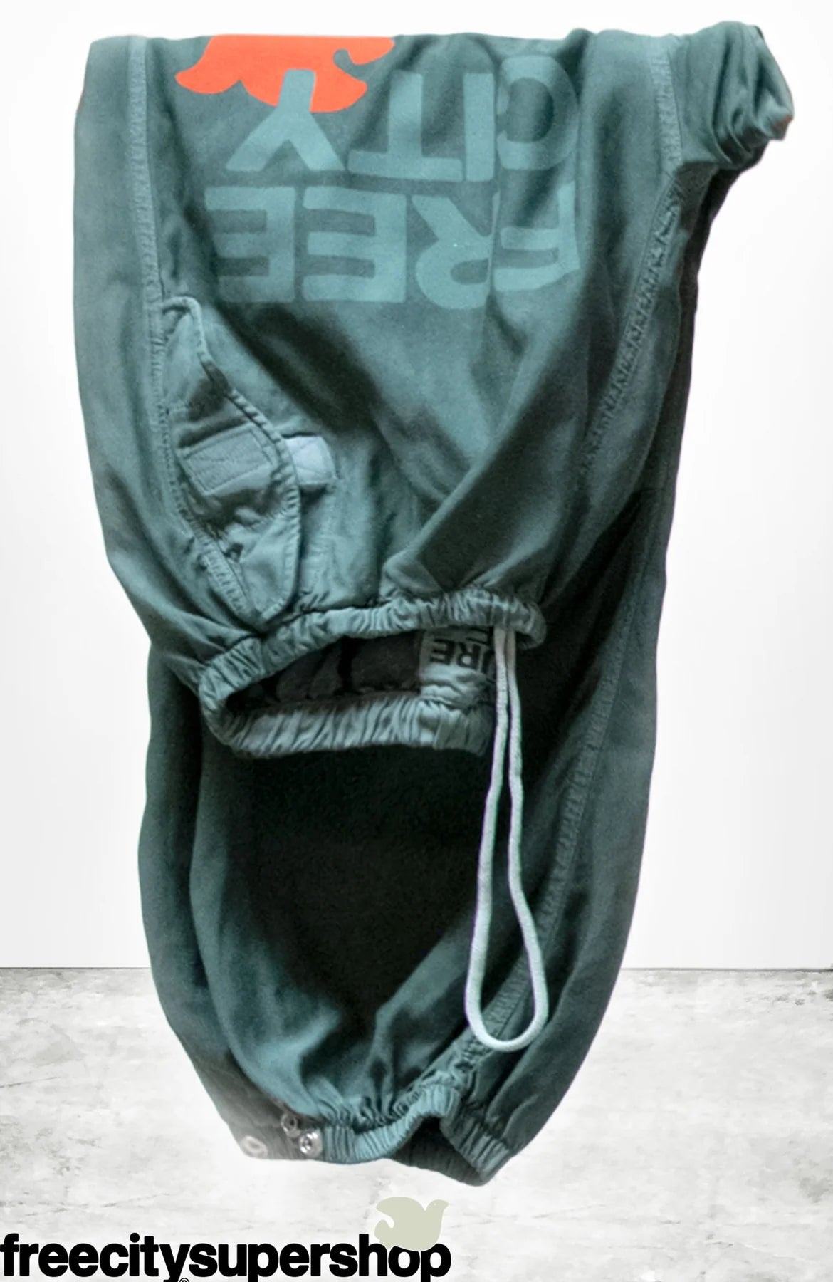 Flap/Snap LG MATTE/SATIN AirJump Pant by Free City (sparrow, LLC) features green fabric with "FREE CITY" and an orange graphic, sunfade washed in Los Angeles. Displayed upside down; "freecitysupershop" shown at the bottom.