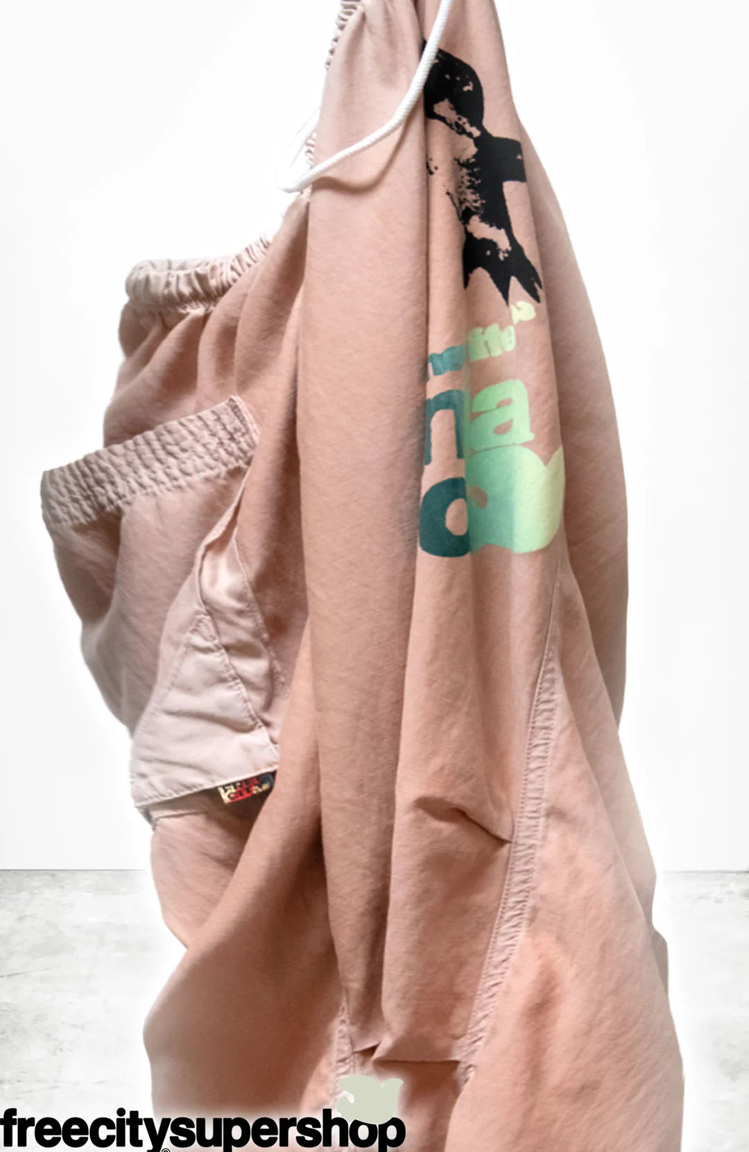 The Loose Drawcord Biggy Pant by Free City (sparrow, LLC) in light pink with graphic print and elastic waistband is displayed against a white background. "freecitysupershop" is shown at the bottom left. Handmade in Los Angeles.