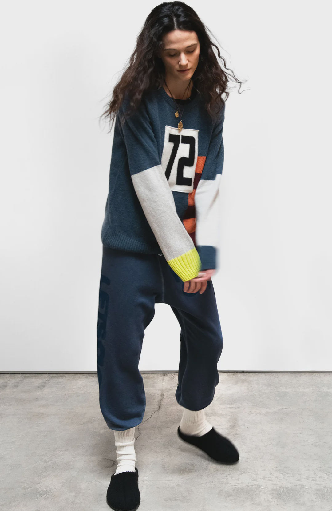 A person with long dark hair wears the Free City (sparrow, LLC) CanyonKid 72' Cashmere Lux Crew sweater, loose blue sweatpants, white socks, and black slippers while standing indoors on a concrete floor against a plain background.