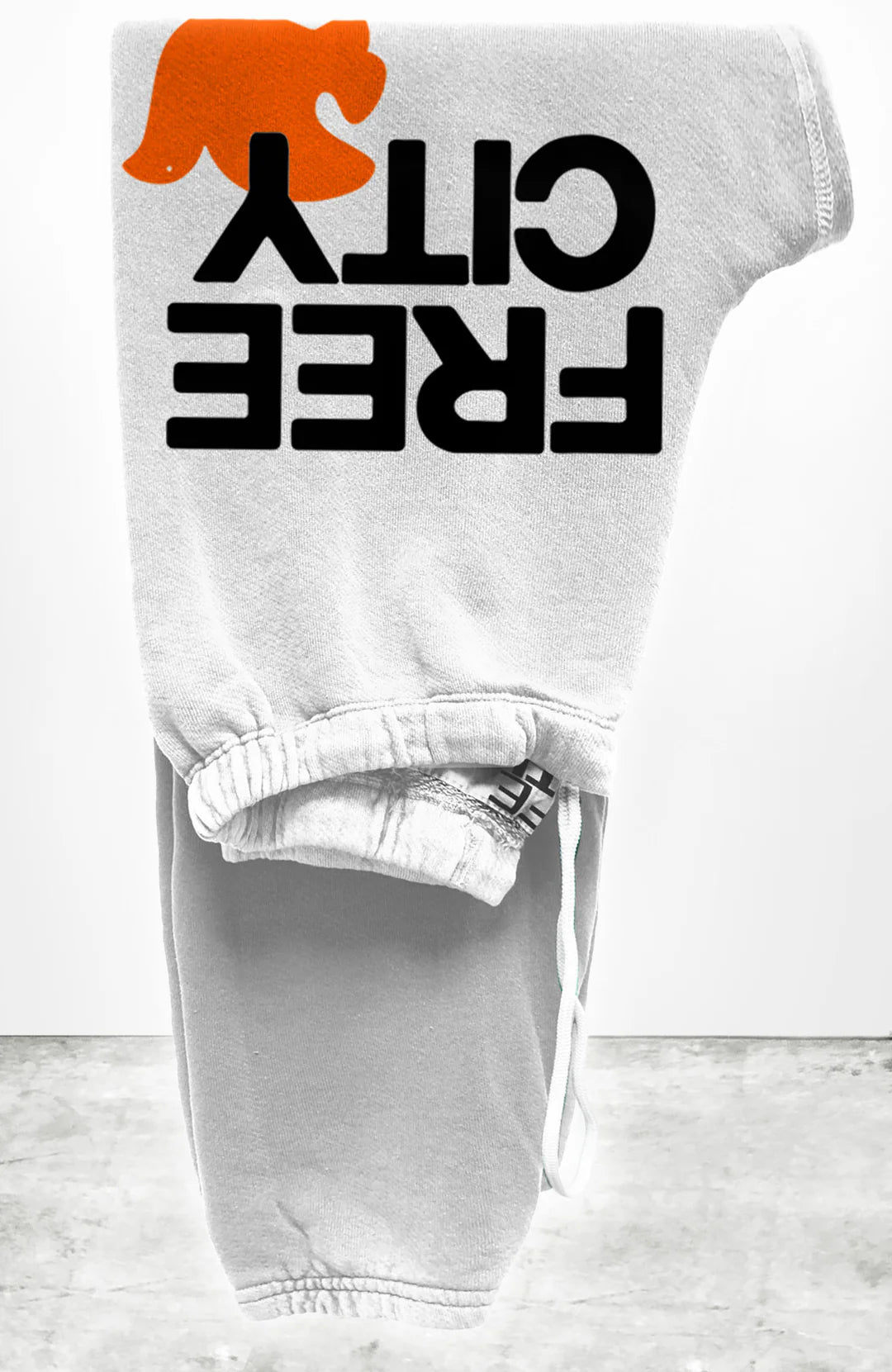 The FREECITY Large Sweatpant by Free City (sparrow, LLC) is a unisex light gray French terry sweatpant, folded neatly with "FREE CITY" in bold black letters and an orange running animal silhouette above the text.