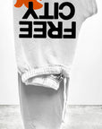 The FREECITY Large Sweatpant by Free City (sparrow, LLC) is a unisex light gray French terry sweatpant, folded neatly with "FREE CITY" in bold black letters and an orange running animal silhouette above the text.