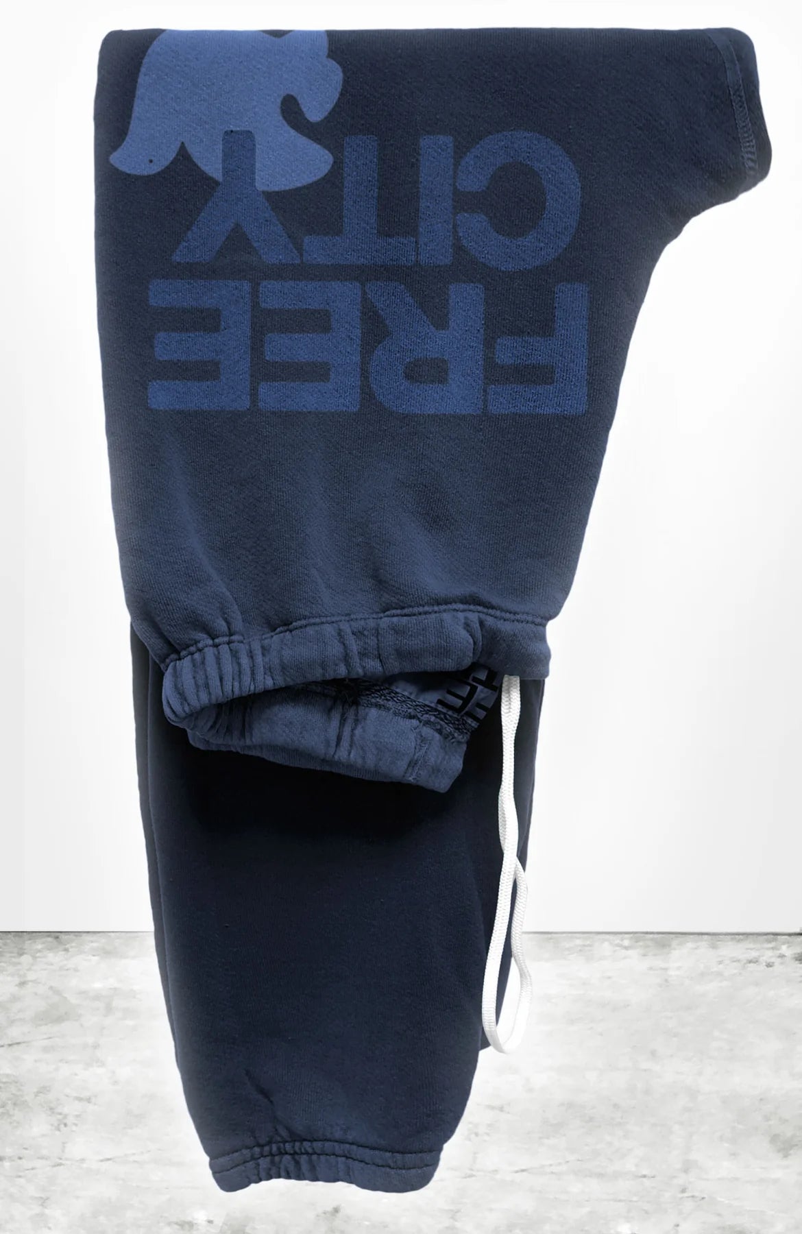 The FREECITY Large Sweatpant by Free City (sparrow, LLC) is a navy blue unisex sweatpant made from soft French terry, featuring a white drawstring and a blue "FREE CITY" graphic with bird logo near the waistband. Seen folded against a white wall and gray floor.