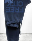 The FREECITY Large Sweatpant by Free City (sparrow, LLC) is a navy blue unisex sweatpant made from soft French terry, featuring a white drawstring and a blue "FREE CITY" graphic with bird logo near the waistband. Seen folded against a white wall and gray floor.