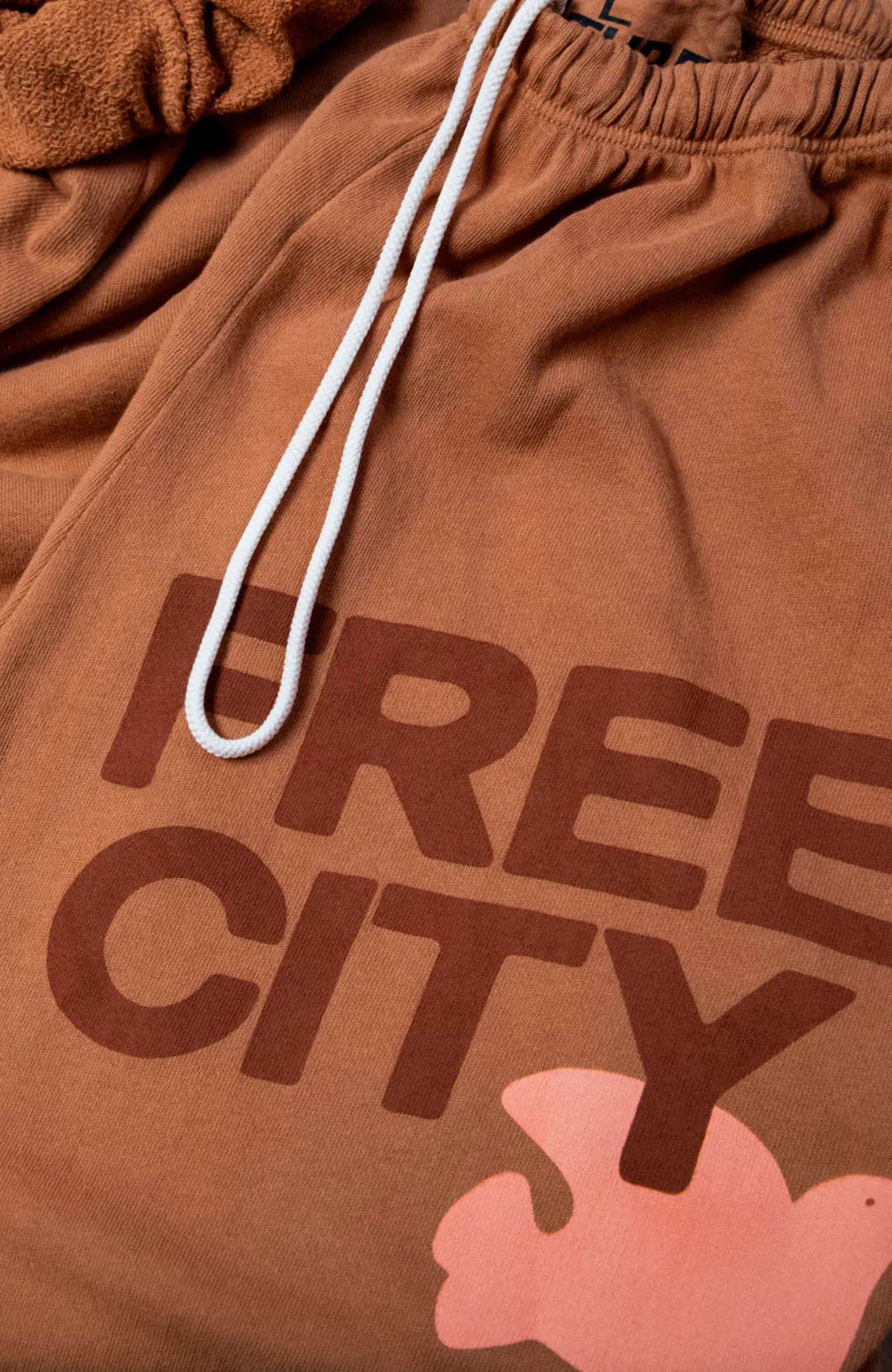 Close-up of FREECITY Large Sweatpant by Free City (sparrow, LLC) in brown French terry with white drawstrings. Features bold “FREE CITY” print and partial pink graphic near the hem. Made in USA for comfort and style.
