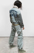 A person with wavy dark hair wears Free City's Flap/Snap AirJump Pant in light gray with a custom sunfade wash and "ARTISTS WANTED" on the back, green high-top sneakers, and a white scarf, standing on concrete facing away from the camera.