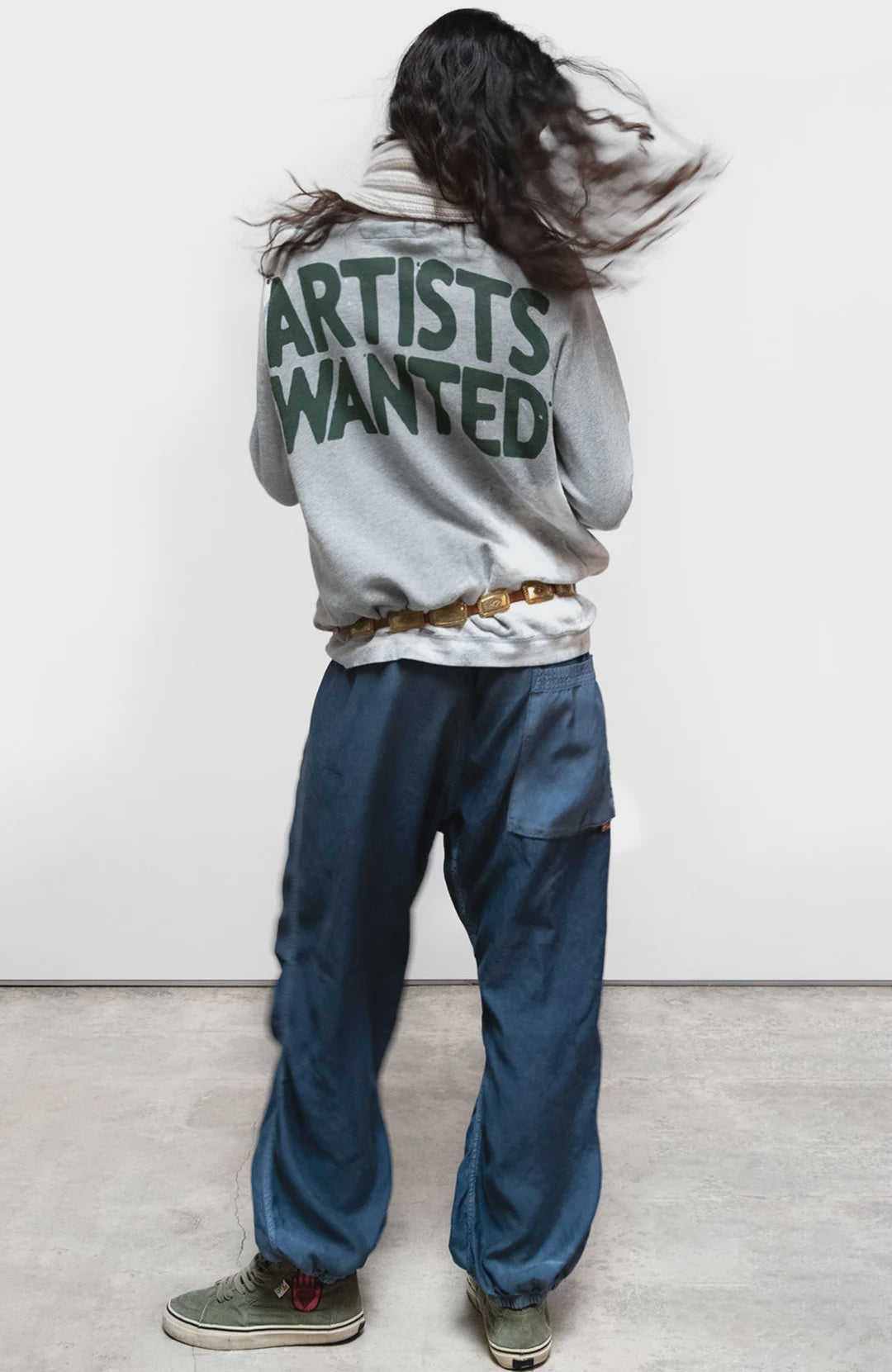 A person with long dark hair, seen from behind, wears the Free City (sparrow, LLC) A. W. Heather Bleachout Sweatshirt with bold green "ARTISTS WANTED" text, loose blue pants, and green shoes, standing on a light concrete floor.