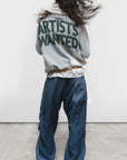 A person with long dark hair, seen from behind, wears the Free City (sparrow, LLC) A. W. Heather Bleachout Sweatshirt with bold green "ARTISTS WANTED" text, loose blue pants, and green shoes, standing on a light concrete floor.
