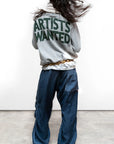 A person with long hair faces away, wearing a gray "ARTISTS WANTED" sweatshirt, Free City Loose Drawcord Biggy Pant by sparrow, LLC, and green sneakers. Set against a plain light background—handmade Los Angeles style.