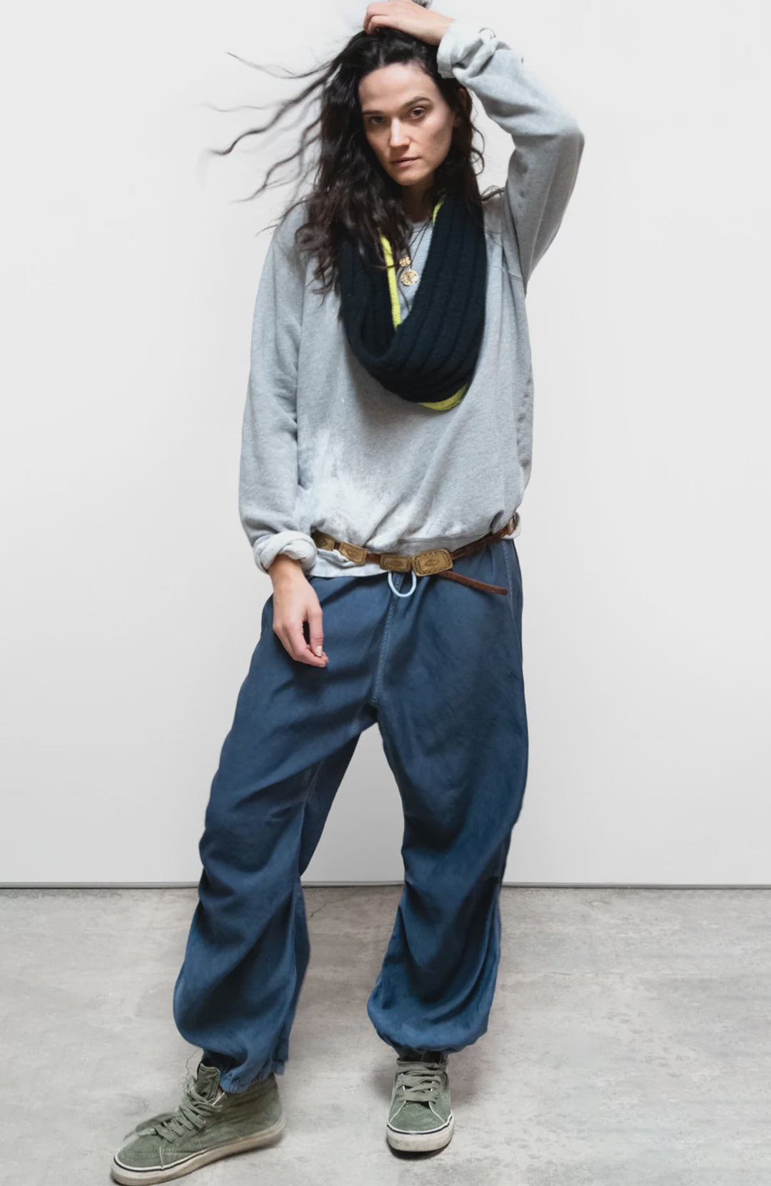 A woman with long dark hair poses against a plain background, wearing Free City Loose Drawcord Biggy Pant, a loose grey sweatshirt, black scarf, and green sneakers. She touches her head as she looks at the camera.