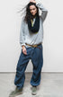 A person poses confidently against a plain background, wearing a loose gray sweatshirt, Free City (sparrow, LLC) Matte/Satin Biggy Pant in an oversized fit, olive green high-top sneakers, a belt, and has dark wavy hair framing their face.