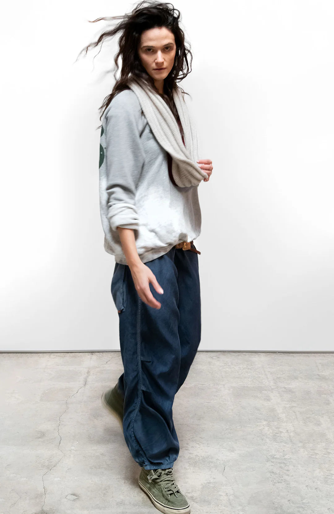 A person with long wavy hair, in a Free City (sparrow, LLC) A. W. Heather Bleachout Sweatshirt, beige scarf, loose blue pants, and green sneakers walks indoors on a concrete floor against a plain white wall.