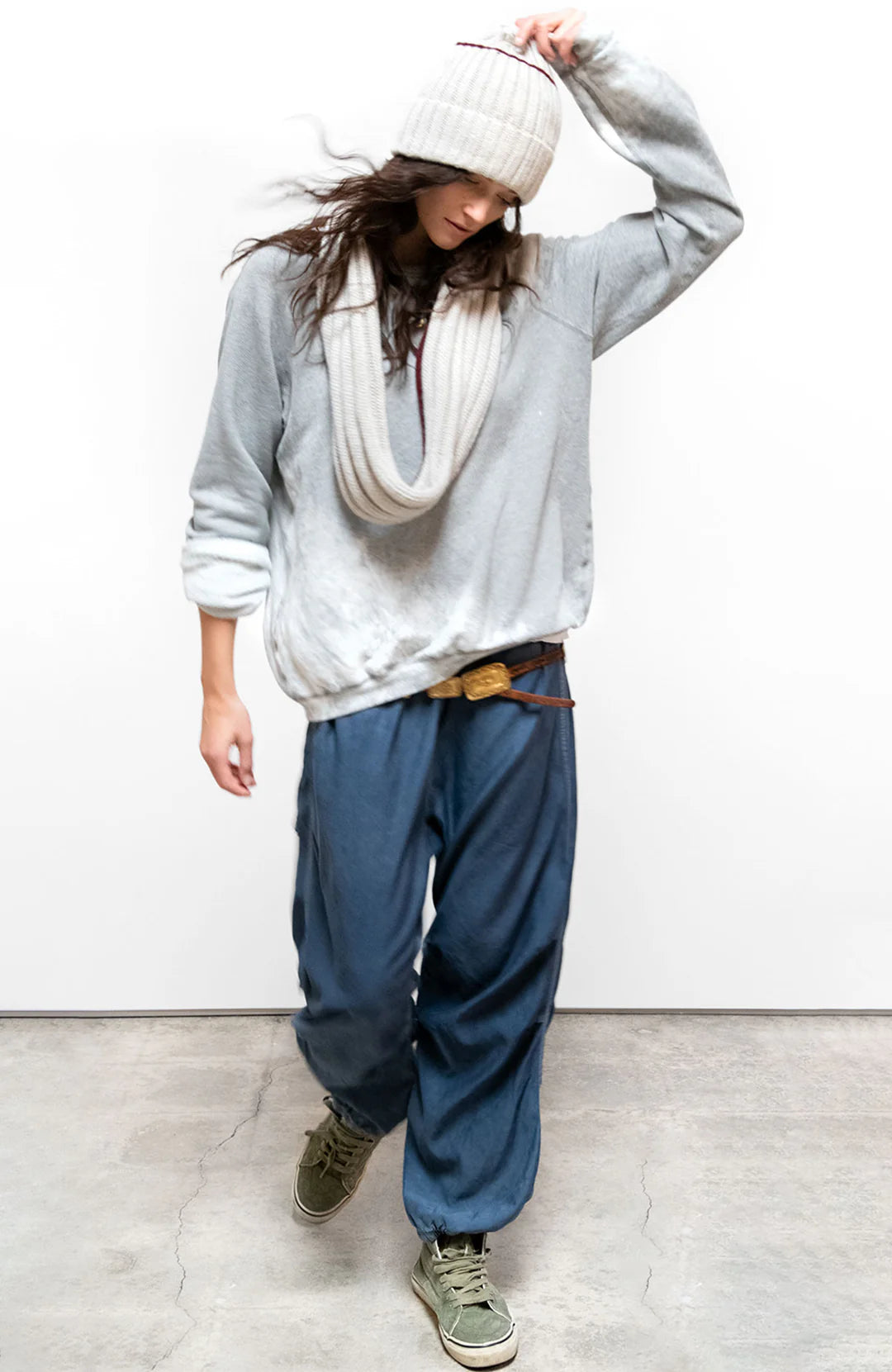 Wearing the Free City (sparrow, LLC) A. W. Heather Bleachout Sweatshirt, a person stands on a concrete floor in a loose gray sweater, scarf, blue baggy pants, and green sneakers, looking down while adjusting their hat.