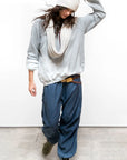 Wearing the Free City (sparrow, LLC) A. W. Heather Bleachout Sweatshirt, a person stands on a concrete floor in a loose gray sweater, scarf, blue baggy pants, and green sneakers, looking down while adjusting their hat.