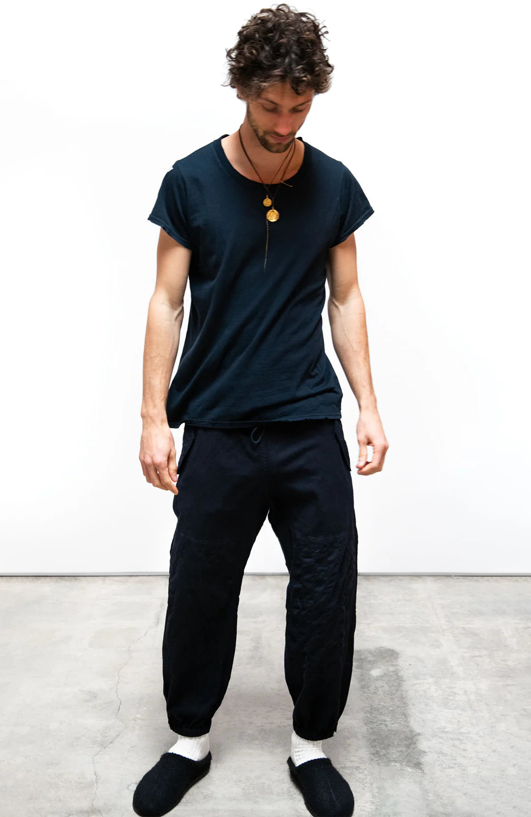 A person with curly hair stands on a concrete floor, looking down. They wear a loose black unisex T-shirt, Free City Cashmere/Wool Flap/Snap Pant, black socks, two gold necklaces, against a plain white background.