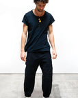 A person with curly hair stands on a concrete floor, looking down. They wear a loose black unisex T-shirt, Free City Cashmere/Wool Flap/Snap Pant, black socks, two gold necklaces, against a plain white background.